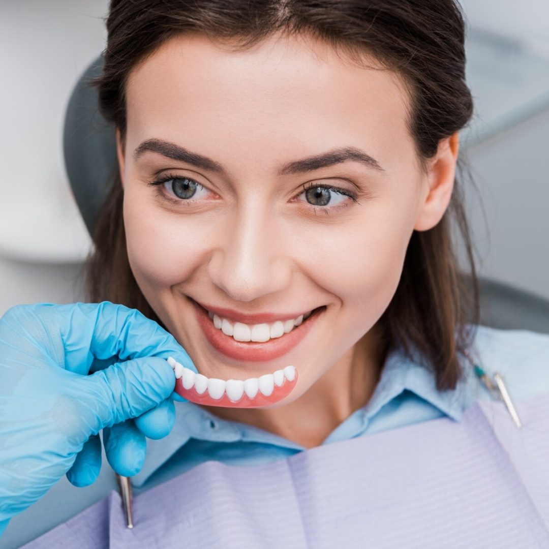 cropped view of dentist holding prosthesis near happy girl in dental clinic